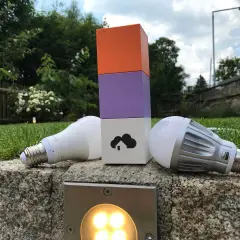 Zwei Glühbirnen liegen neben einem bunten Turm, der die Wahl zwischen Z-Wave vs. Zigbee symbolisiert.