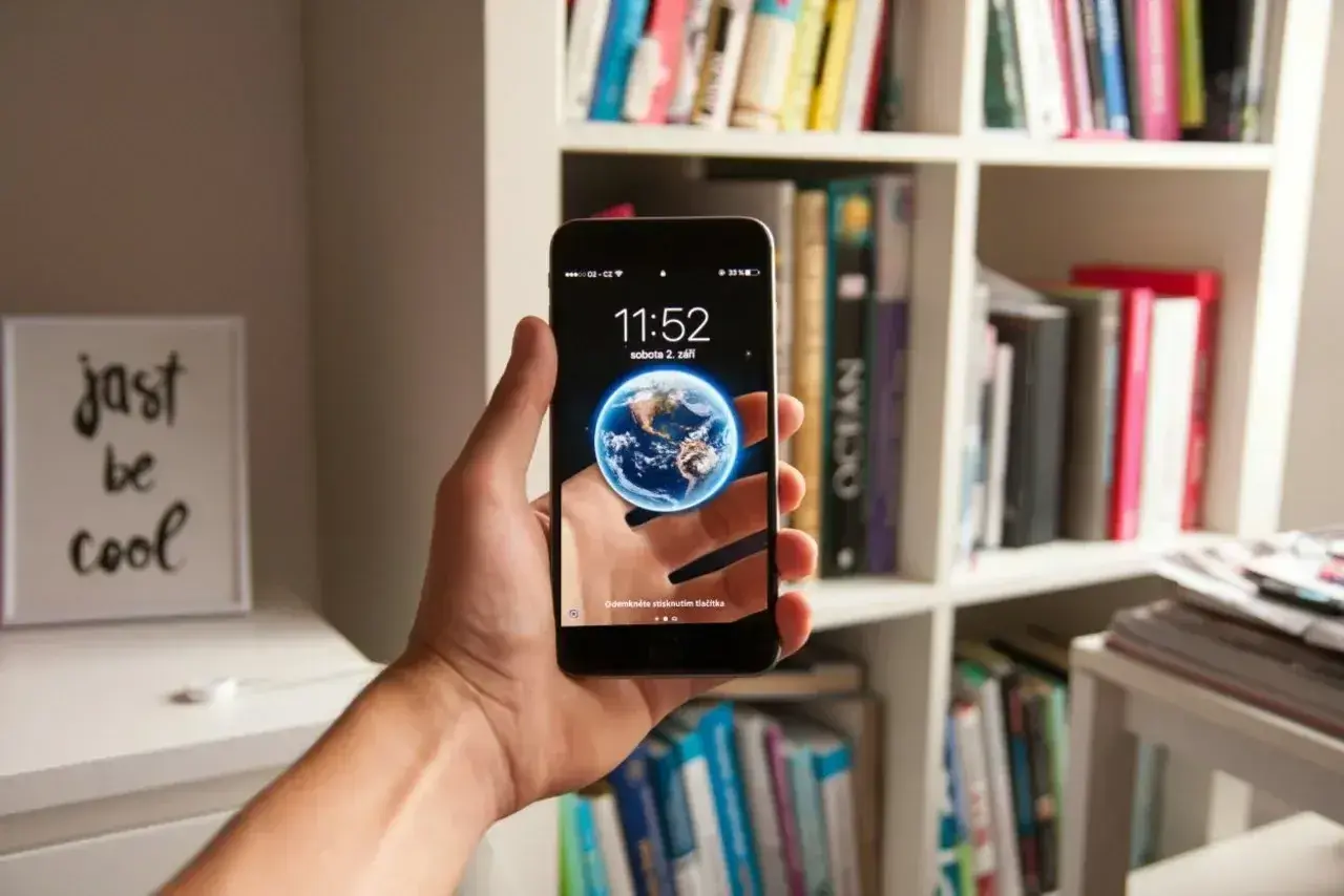 Hand hält Smartphone mit Erd-Hintergrund. Das neue Betriebssystem zeigt die Uhrzeit und das Datum. Im Hintergrund ein Bücherregal.
