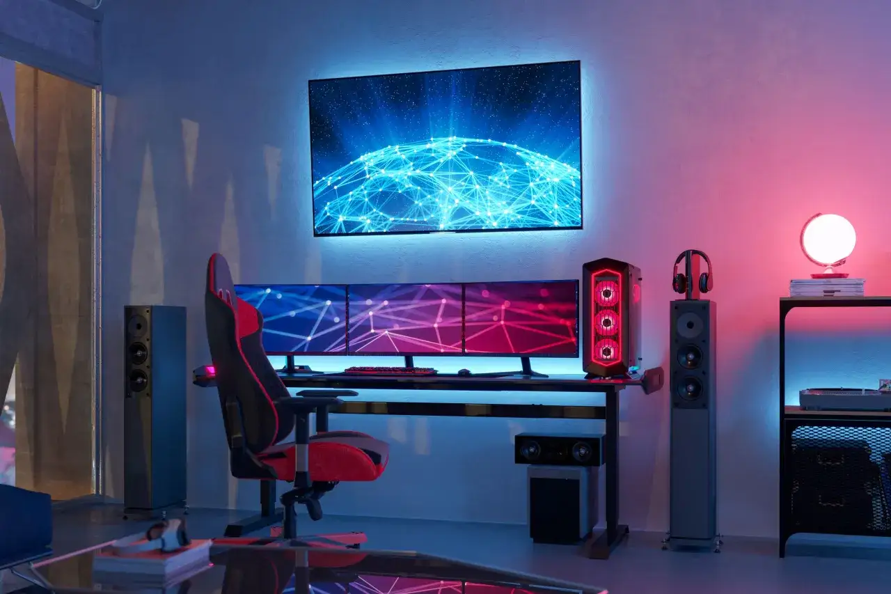 Gaming-Setup mit drei Monitoren, die getrennt genutzt werden k&ouml;nnen. Ein gro&szlig;er Bildschirm h&auml;ngt dar&uuml;ber. Roter Gaming-Stuhl, PC und Lautsprecher.