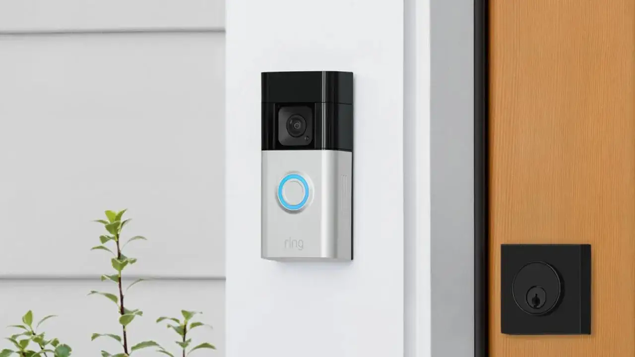 Ring Video Doorbell: Ein silbernes Ger&auml;t mit Kamera und blau leuchtendem Knopf an einer wei&szlig;en Wand. Nebenan eine Holzt&uuml;r mit schwarzem Schloss. Vergleiche Ring Doorbell Unterschiede.